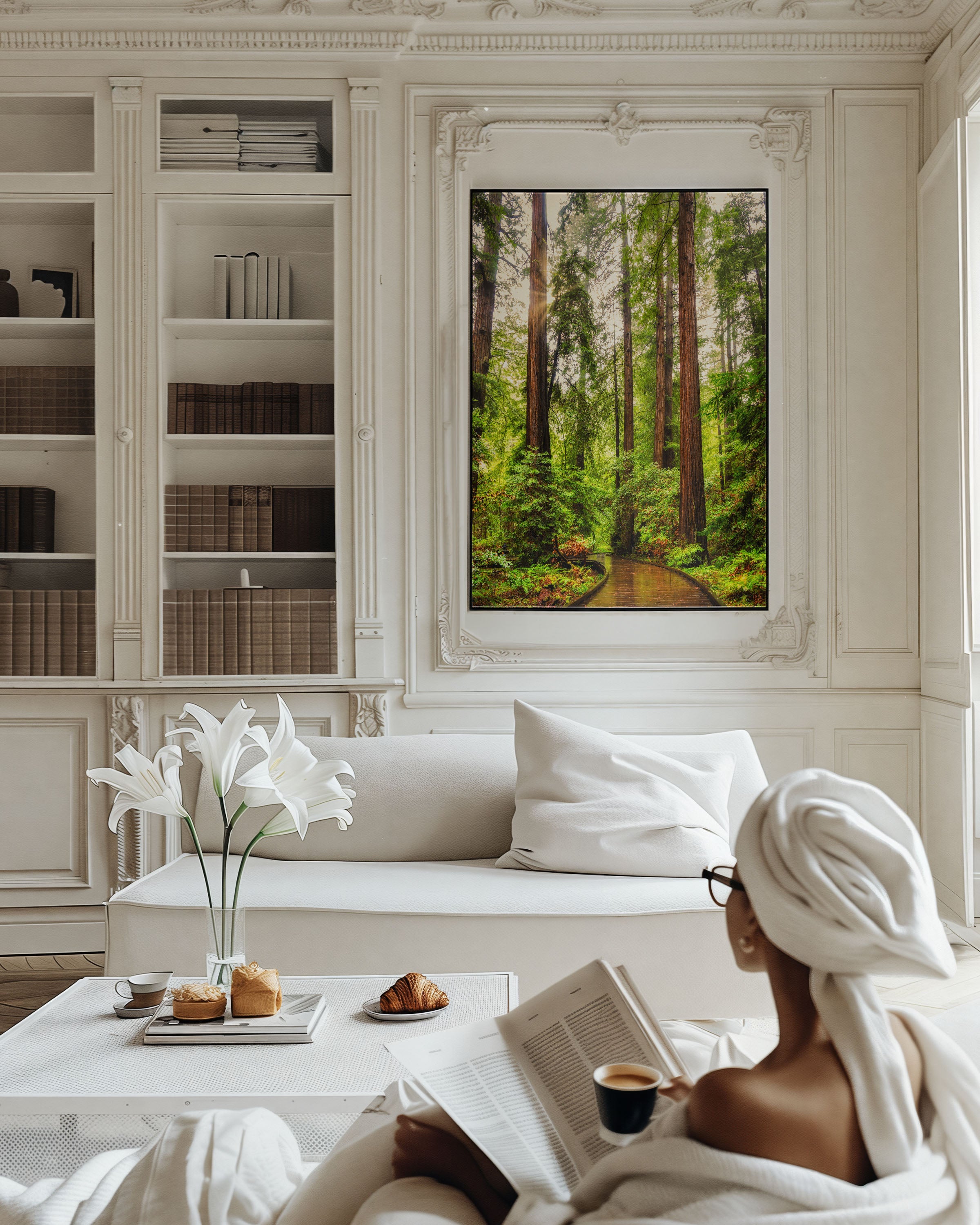 A framed forest landscape print hangs on a white paneled wall above a sofa in a bright, elegant living room. A woman wrapped in a white towel sits on the floor reading a book near a coffee table with flowers and pastries.