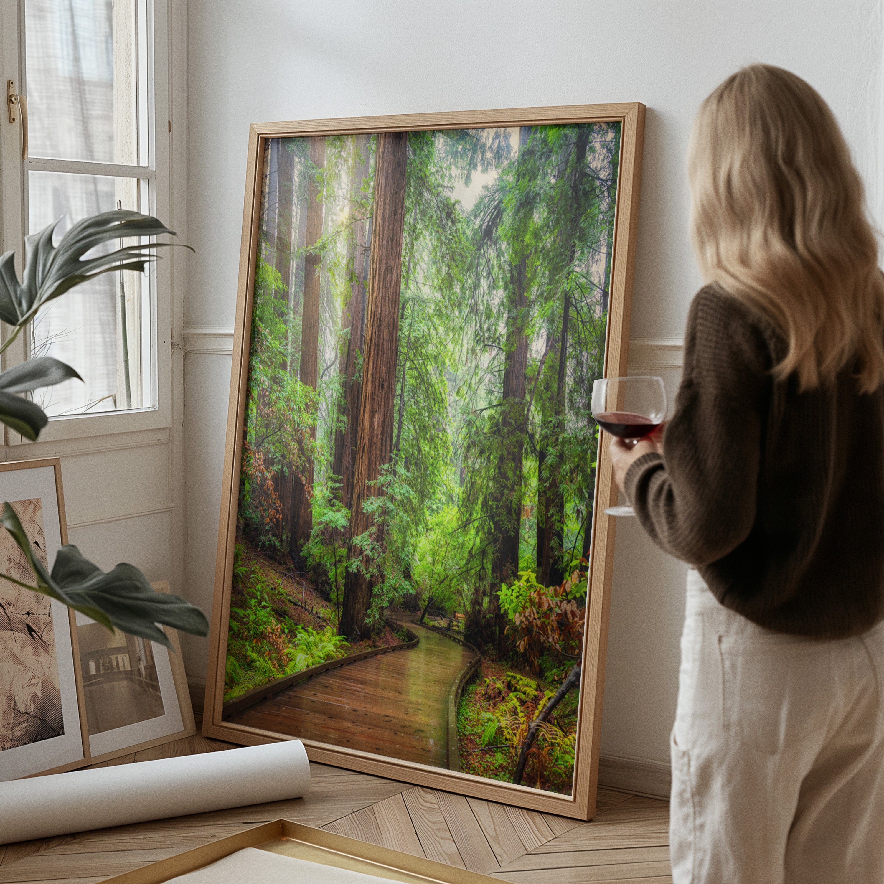 A woman stands beside a large framed redwood forest art print in a light wood frame, holding a glass of red wine in a bright room with a window, plant, and rolled canvas on the floor.