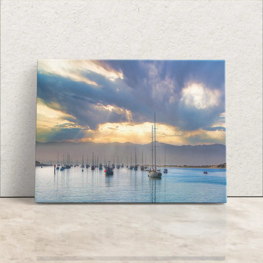A canvas print leaning against a white wall features a serene harbor at sunrise with moored sailboats and dramatic clouds.