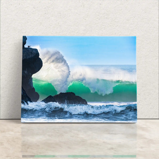 A canvas print of a powerful ocean wave near Morro Rock, leaning against a white wall, showcasing vibrant blues and greens.