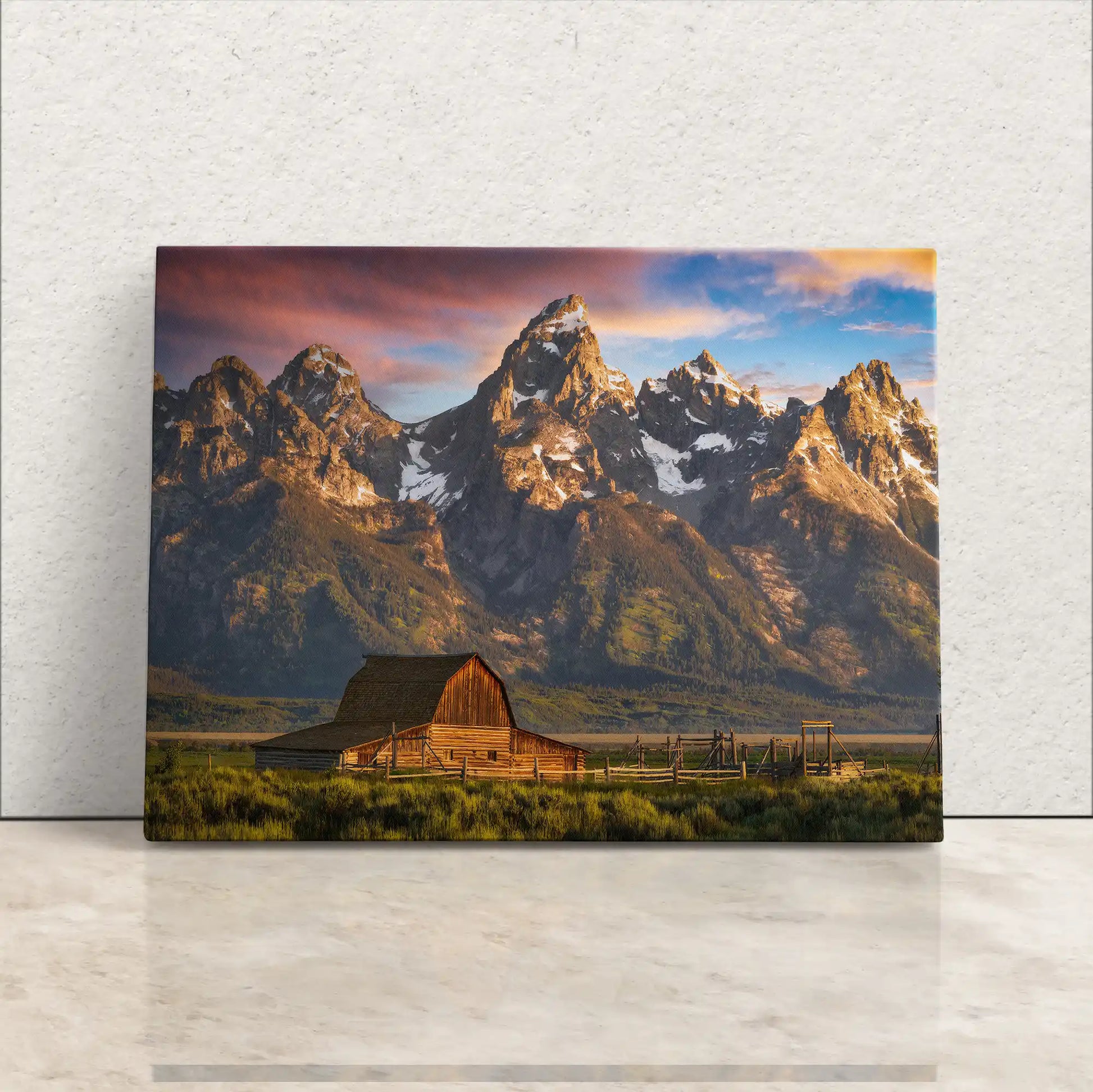 A canvas print leaning against a white wall, depicting the John Moulton Homestead with the Teton Mountains in the background, bathed in the warm light of sunrise.