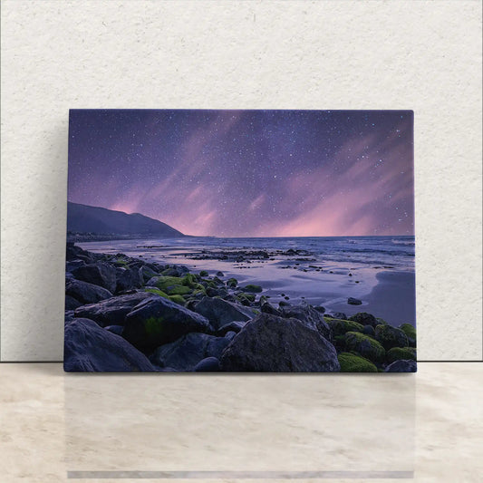 Canvas print of a purple twilight seascape with a starry sky over a beach strewn with rocks and green moss.