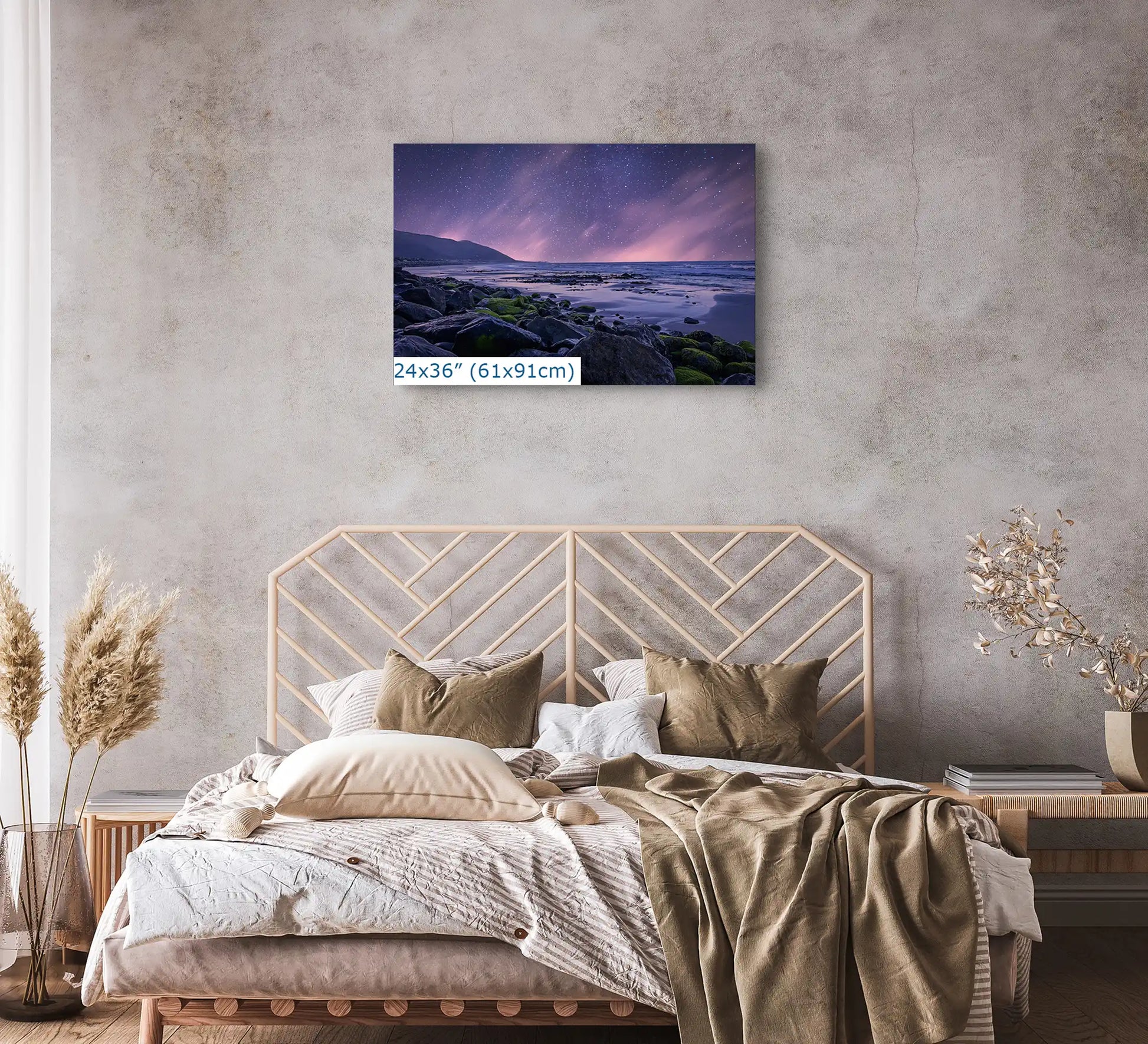 A 24x36 inch canvas of a starry night sky and purple twilight over a rocky beach, placed above a bed.