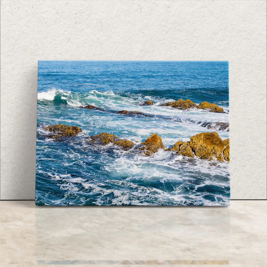 A canvas print leaning against a white wall, depicting a vibrant ocean scene with frothy waves crashing onto sunlit, rugged rocks.