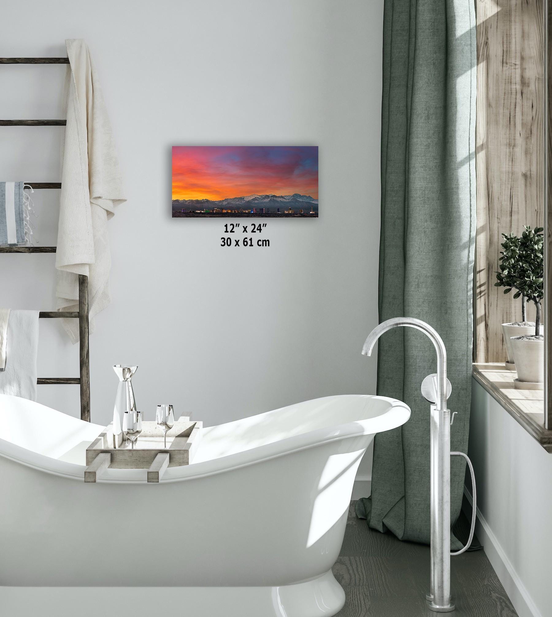 Framed 12x24 metal print of the Las Vegas skyline at sunset hung above a bathtub in a modern bathroom setting.