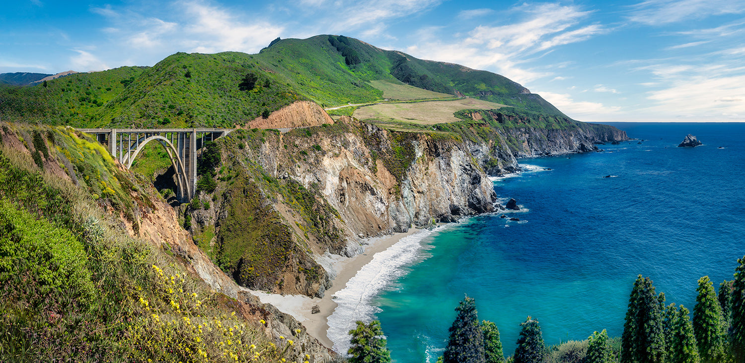 Scenic coastal landscape of Bixby Bridge spanning a cliff over turquoise ocean water, a sandy beach, green hills, and blue sky with clouds.