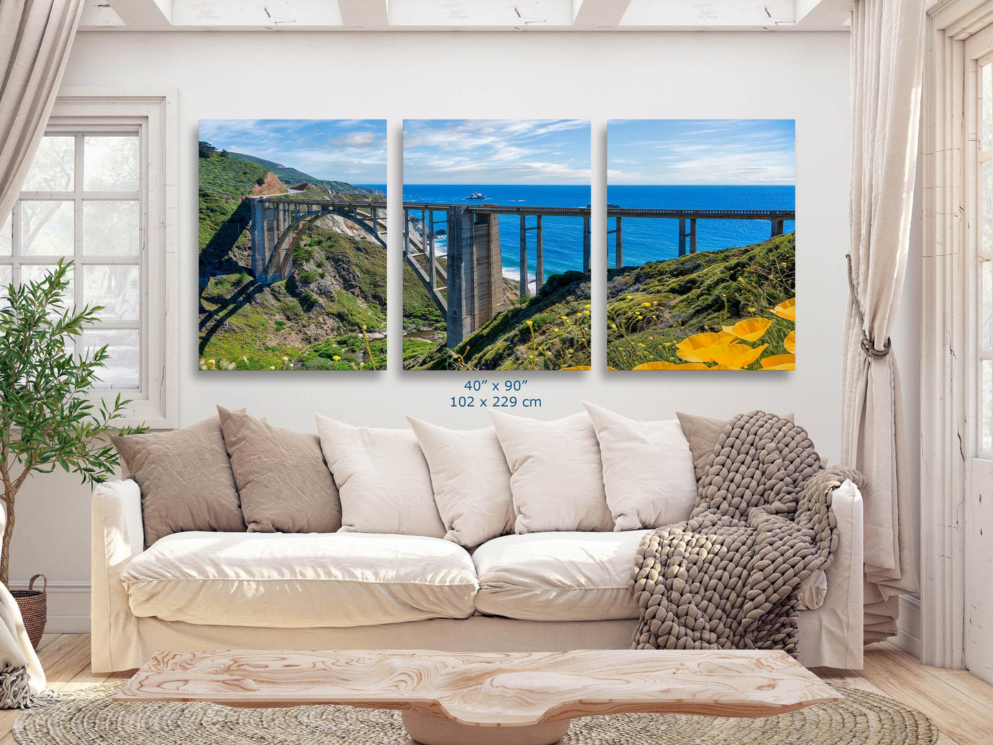 Three-panel triptych wall art featuring Bixby Bridge on the Big Sur coast of California, displayed above a couch in a living room, size 40x90 inches.