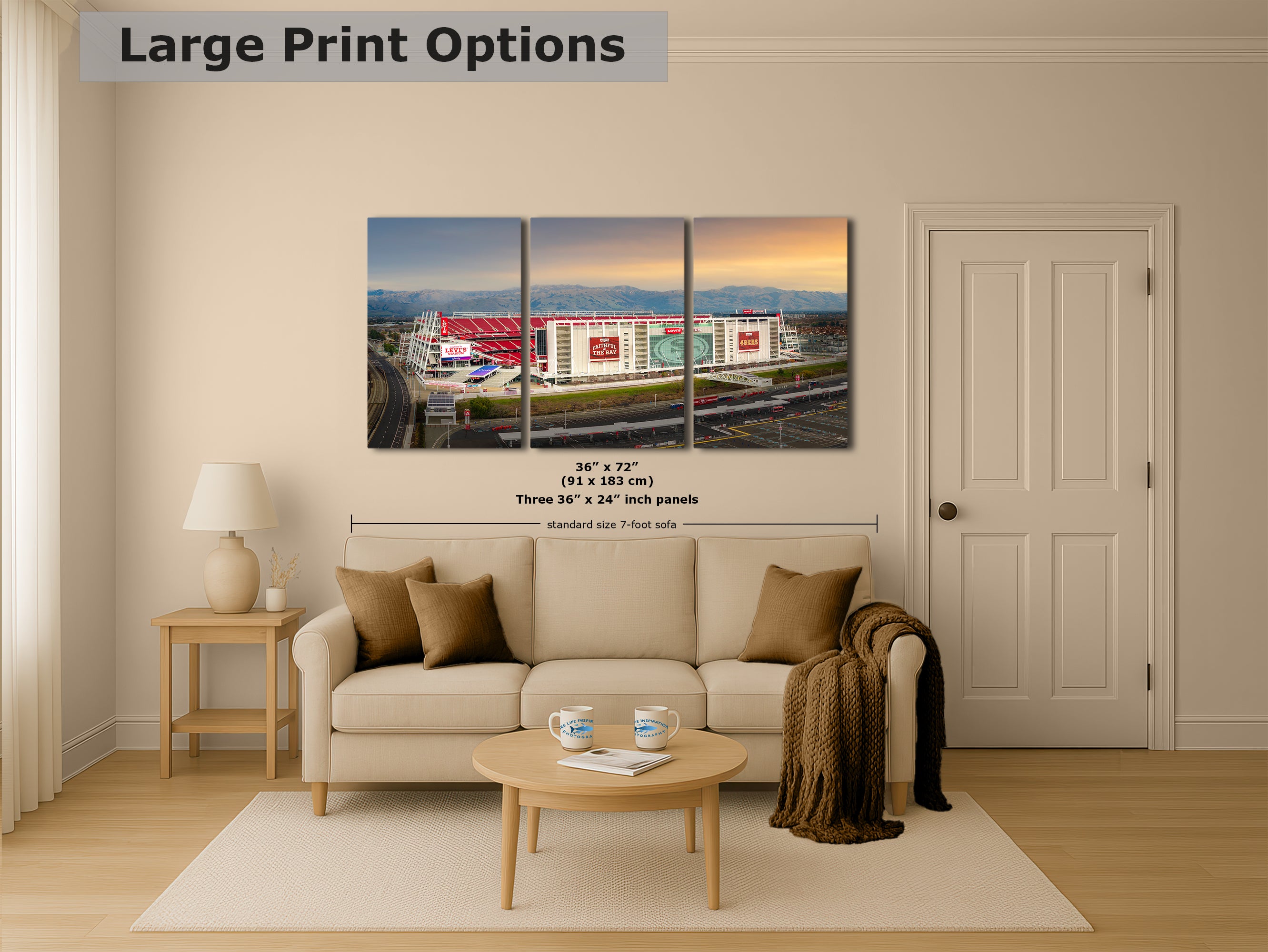 The image is a living room with a large, three-part canvas print depicting a cityscape with a stadium in the background. The print is mounted on the wall above a beige sofa and a coffee table.
