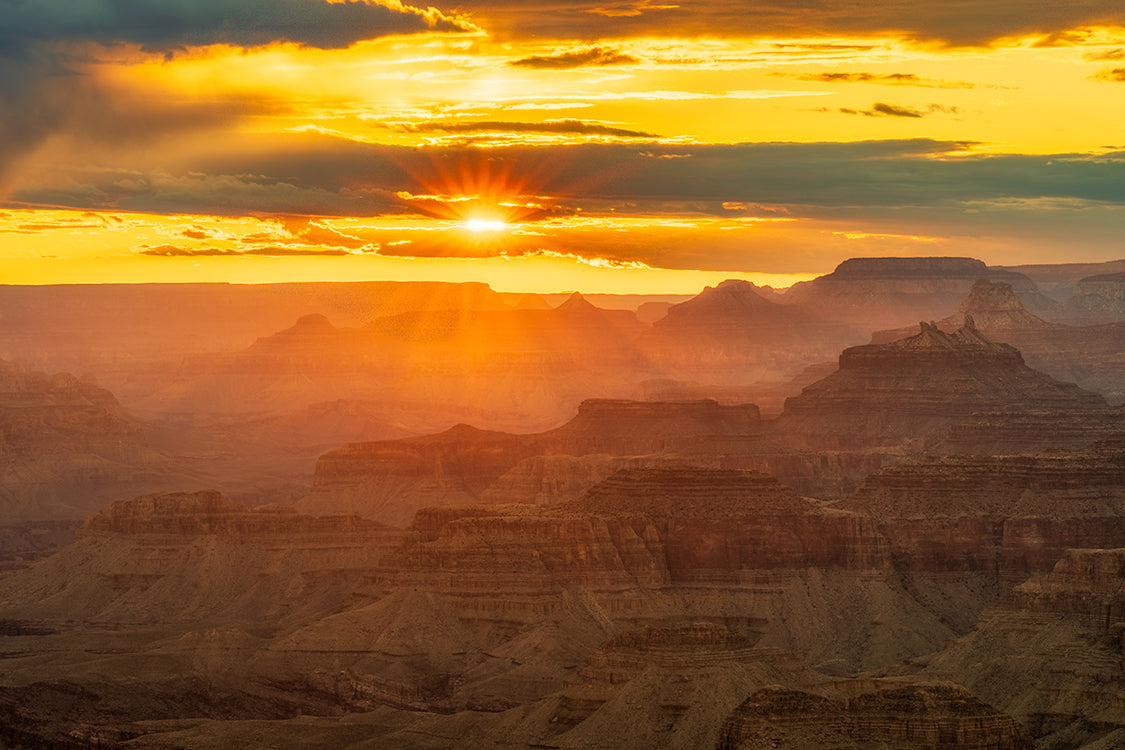 A dramatic sunset over the Grand Canyon, with golden sunlight breaking through clouds and illuminating layered rock formations in warm orange and brown tones.