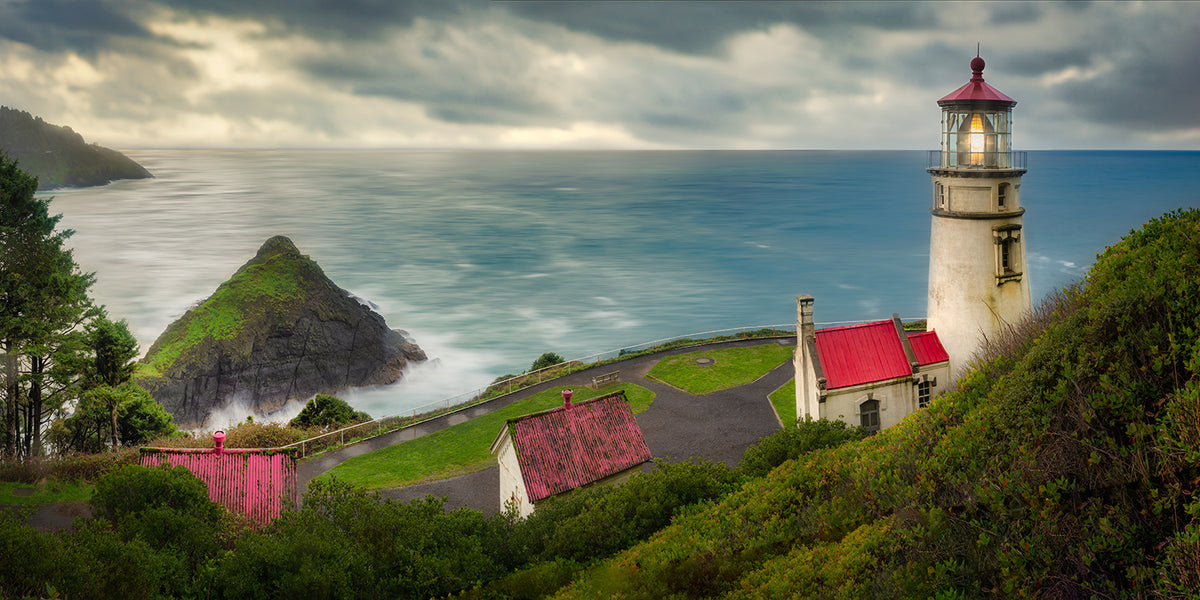 A coastal art print of Heceta Head Lighthouse on a grassy cliff overlooking the ocean, with small buildings featuring red roofs, dramatic storm clouds, and a rocky sea stack in the background.