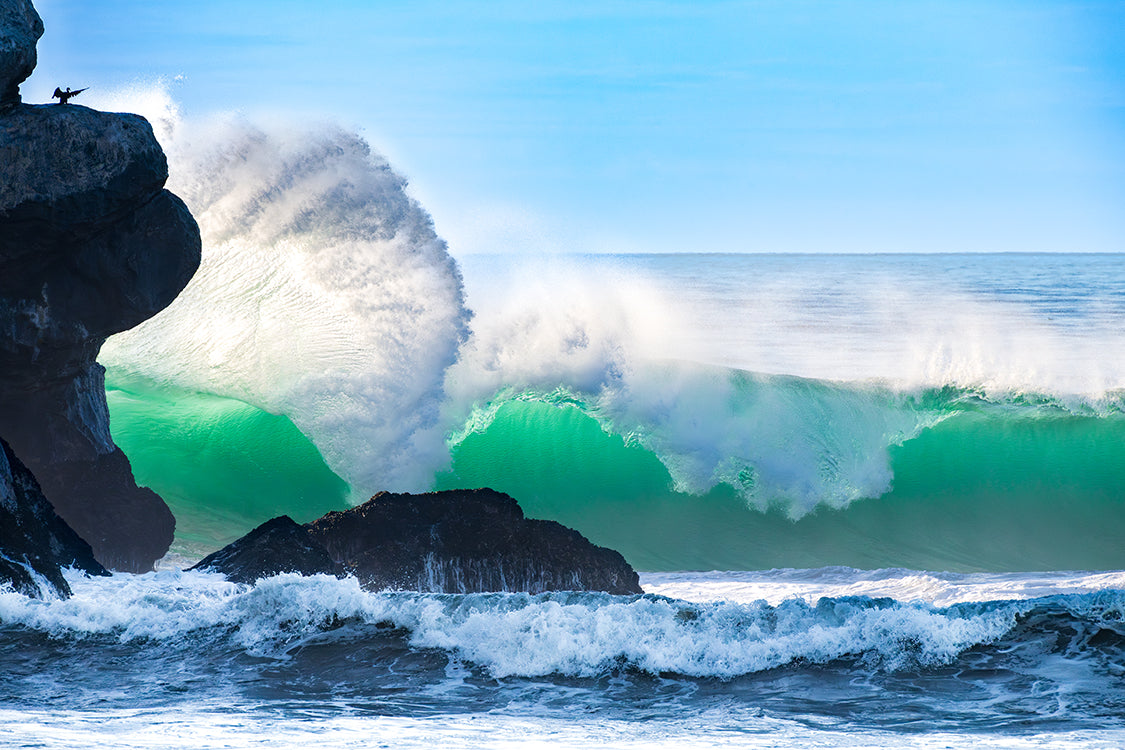 A dramatic ocean wave with bright green water and white foam crashing near dark rocky cliffs under a clear blue sky.