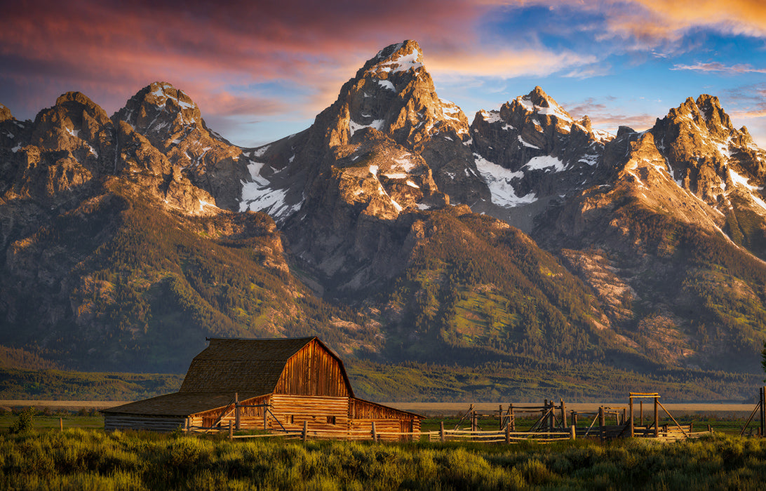A scenic landscape print featuring a rustic wooden Moulton Barn in a grassy field, set against the towering, snow-capped Grand Teton Mountains under a dramatic sunrise sky.