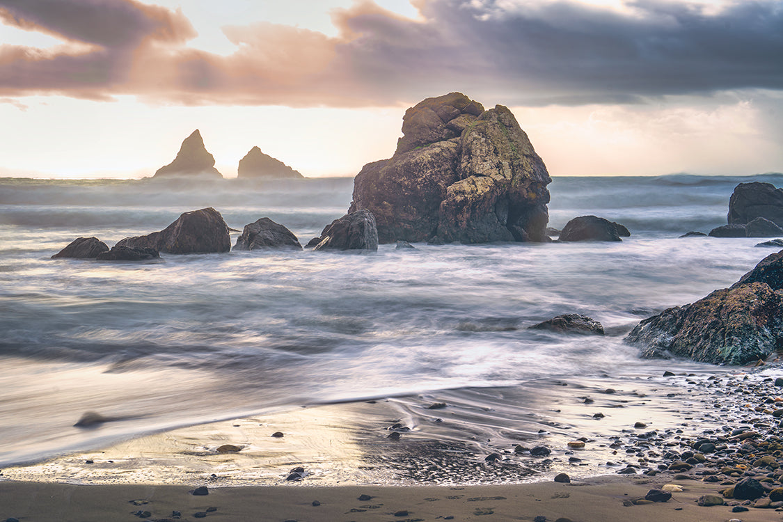 A long-exposure seascape photograph of ocean waves washing onto a sandy beach, with large dark sea stacks and rocks rising from the water under a dramatic cloudy sky at sunset.