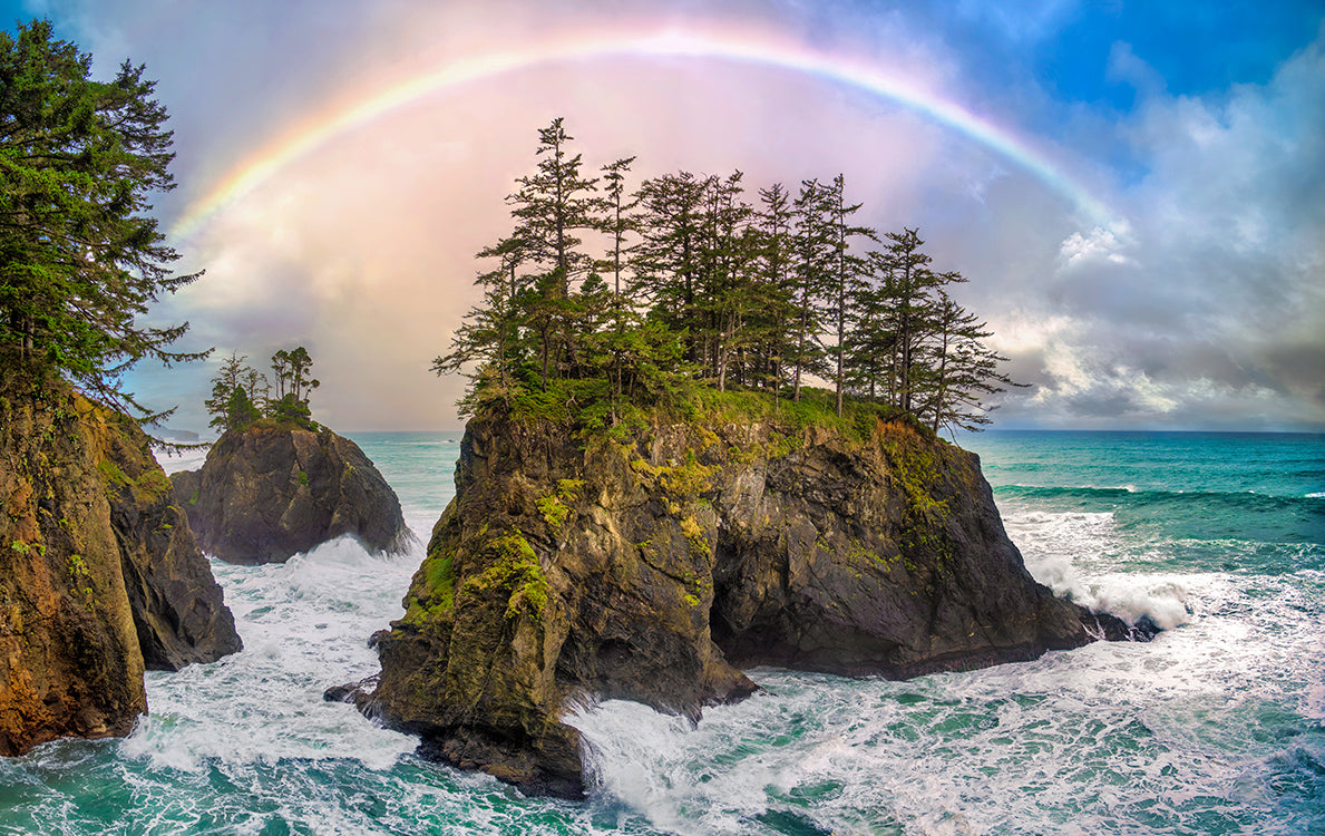 A dramatic coastal landscape with rocky sea stacks rising from turquoise ocean water, green trees on the rocks, and a large rainbow arching across a cloudy blue sky.